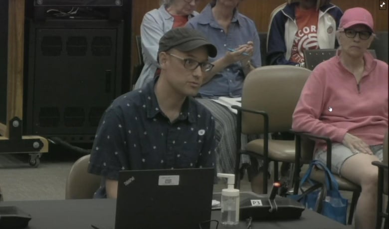 A man wearing a black cap and a blue shirt speaks in a council chamber.