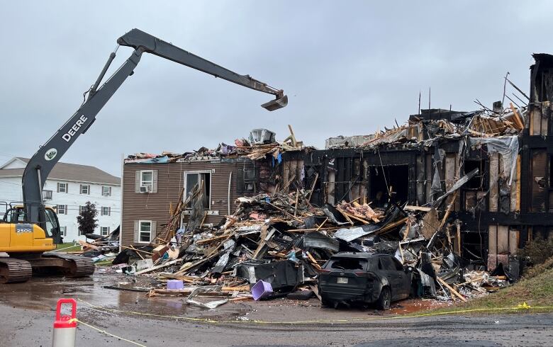 A backhoe tears at a burned out building, with a charred car in the foreground.