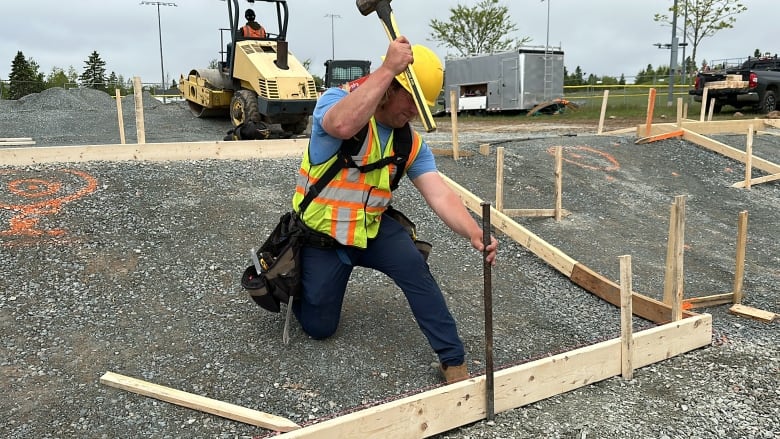 A worker in a yellow and orange safety vest is shown kneeling down hammering a spike into gravel as part of work making a concrete form. 