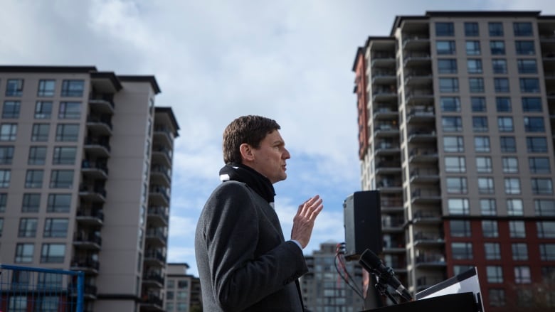 A white man in a dark coat stands in profile with high rises in the background.