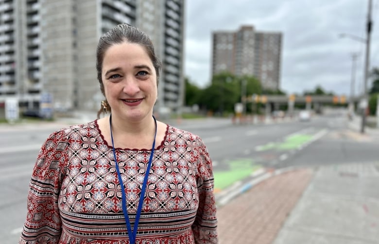 A woman stands looking into the camera, a streetscape behind her