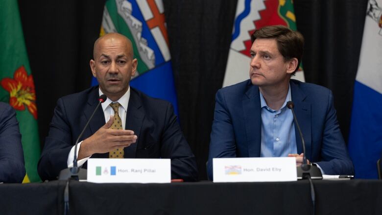 Two men sit side-by-side at a table behind microphones, with a backdrop of flags.