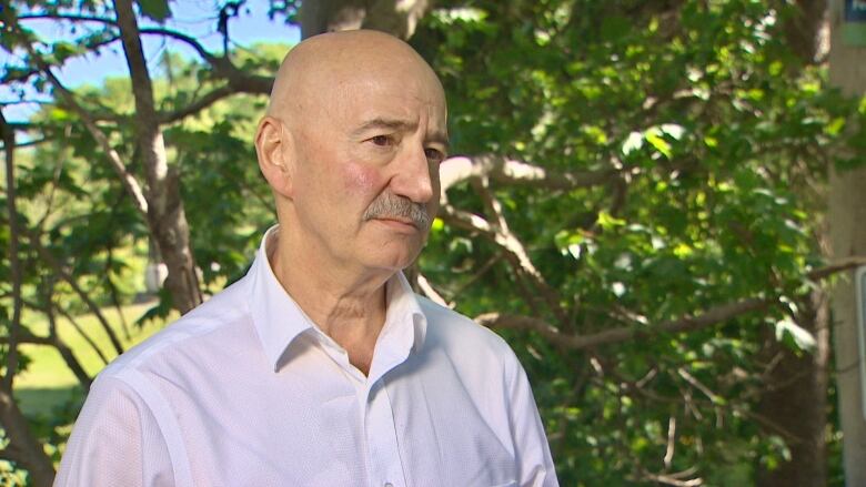 A man in a white dress shirt stands in front of a leafy tree.