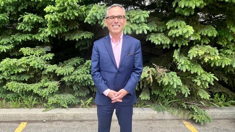 A man in a lue suit stands in front of a row of fir trees.