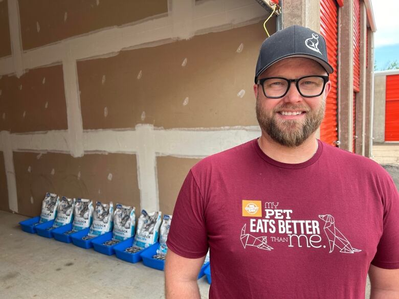 A bearded man with glasses stands beside a storage unit that has bags of cat food and blue plastic litter boxes lined neatly against the wall. 