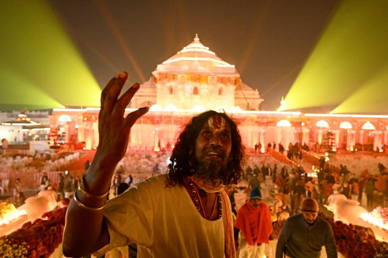 A man dressed in yellow with gold and red jewelry stands with his right hand up. Behind him is an illuminated temple and crowds of people. 