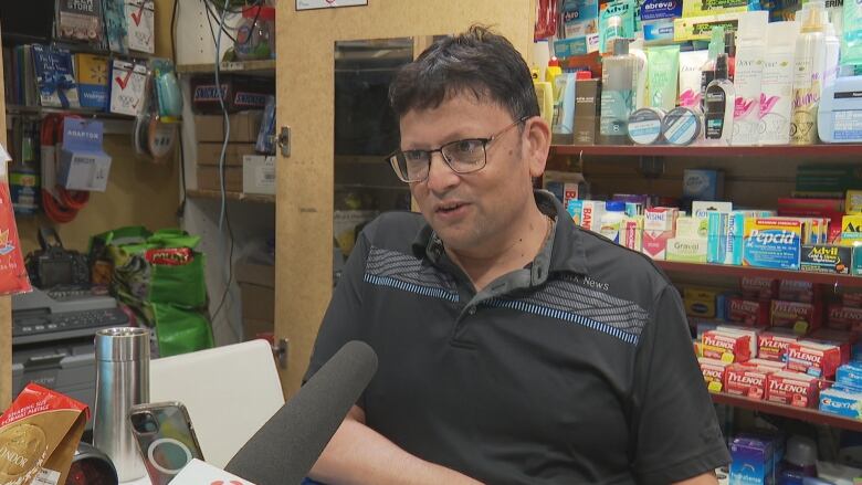 A man behind a corner store counter speaks into a reporter's microphone