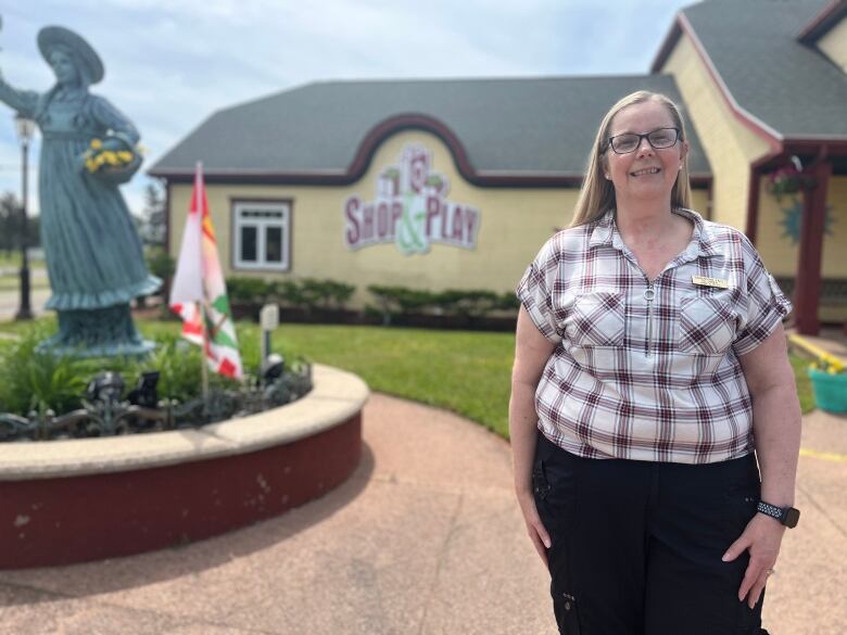 A woman stands outside a yellow building, a gift shop, with a statue of Anne of Green Gables near her. 