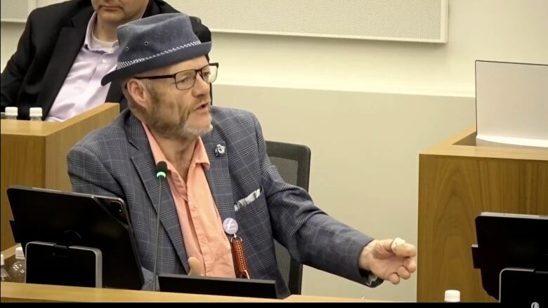 A councillor sits in a council chamber; he's wearing a fedora. 