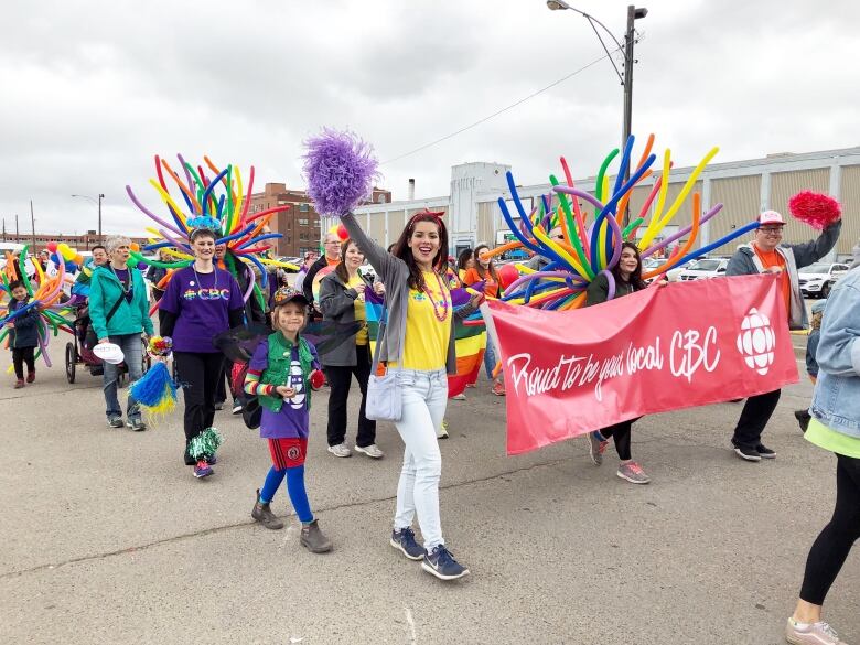 A woman in a yellow shirt with and white jeans smiles while holding a purple pom pom and giant red banner that says Proud to be your local CBC