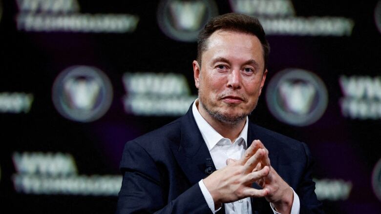 A man in a black suit, white button-down shirt and no tie folds his hands as he speaks at a tech conference.