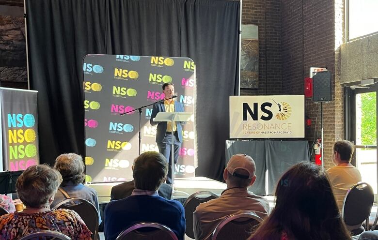 A man stands behind a podium and microphone. People on chairs lined up in front of him are looking at him. There are several posters reading 