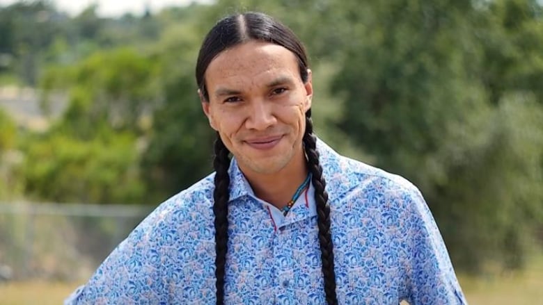 a man smiles to camera wearing a blue shirt and long brown braids in his hair with green trees in the background