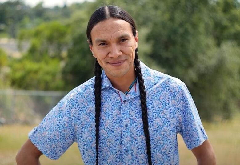 a man smiles to camera wearing a blue shirt and long brown braids in his hair with green trees in the background