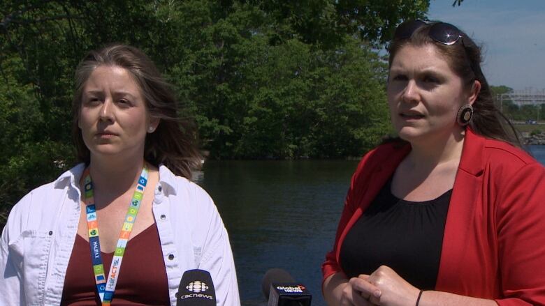 Two women stand in front of a lake with leafy green trees in the back. One wears a white jacket, the other wears a red blazer. They both have brown hair. 