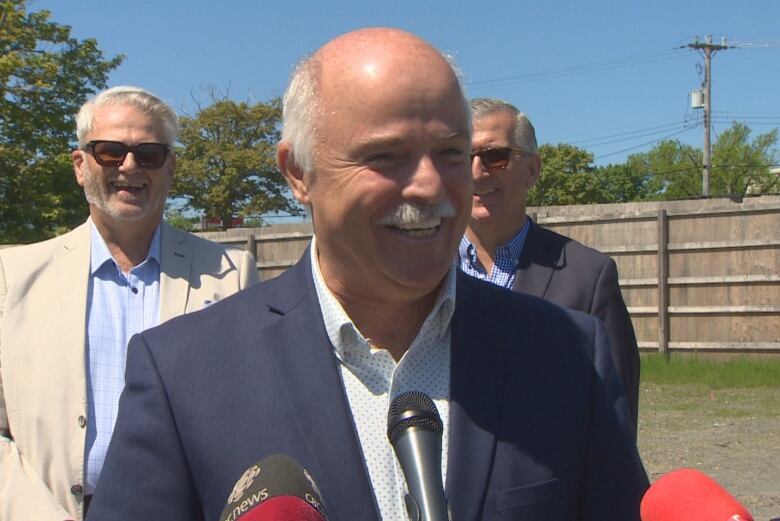 A smiling man in a dark suit stands outside in front of a trio of microphones.