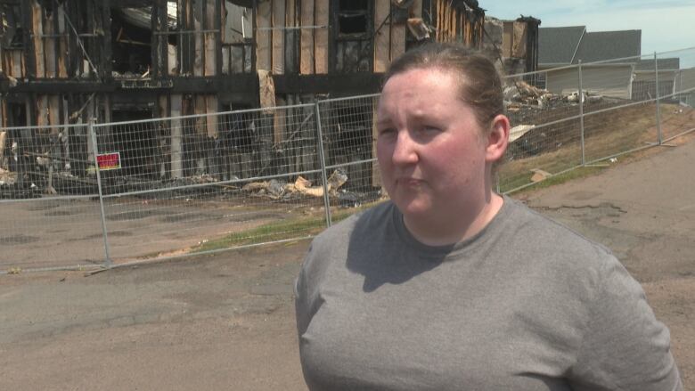 A woman with dark hair in a ponytail and a plain grey t-shirt standing in front of a burned down apartment complex.