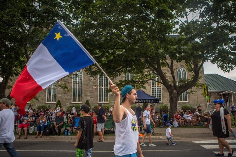 A man wearing a tank top and a baseball hat waves an Acadian flag.