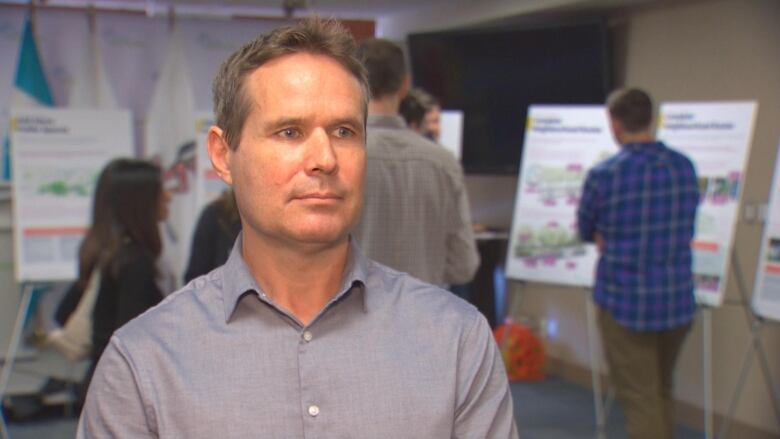 A white man wearing a button-up grey shirt looks at the camera in a room surrounded by charts and boards.