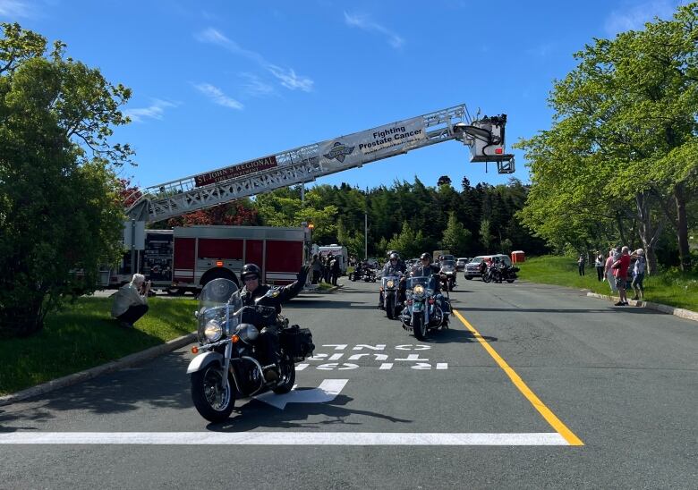 People on motorcycles are driving down a road. The man on the motorcycle in the foreground waves. A fire truck is parked in the background. Its ladder is extended and a banner attached to it reads 