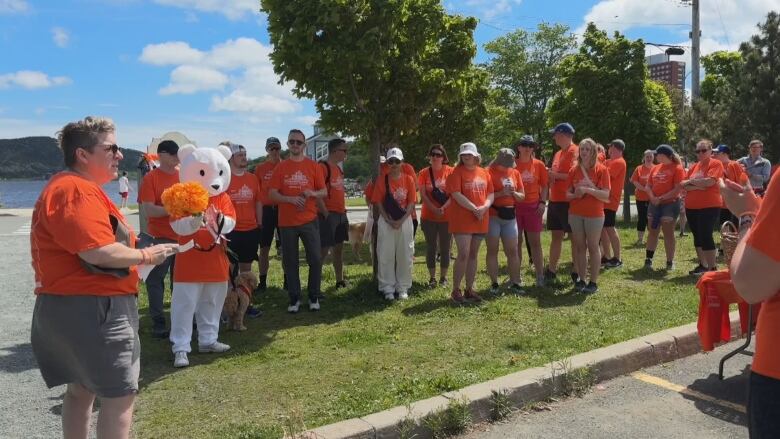 Several people in orange shirts are gathered in a semi-circle. A bear mascot is also in the crowd. In the distance, a body of water is visible. 