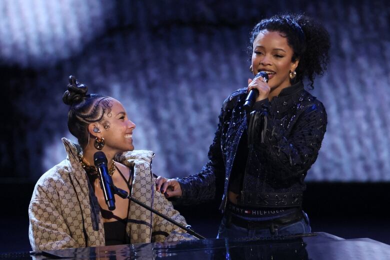 A woman seated at a piano smiles at a woman standing beside her singing and holding a microphone.