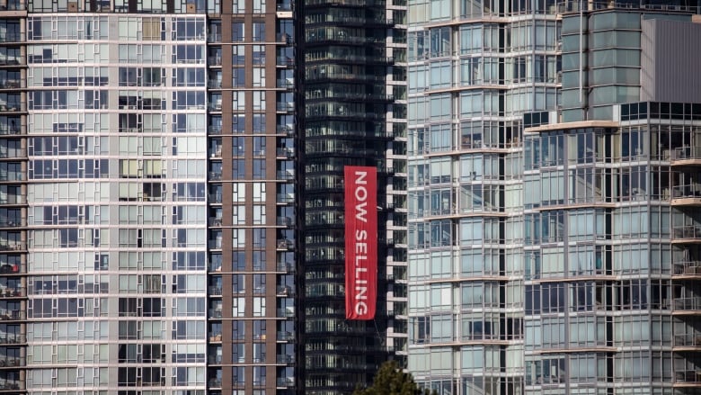 A red banner printed with the words 'now selling' hangs from a condo in a downtown setting.