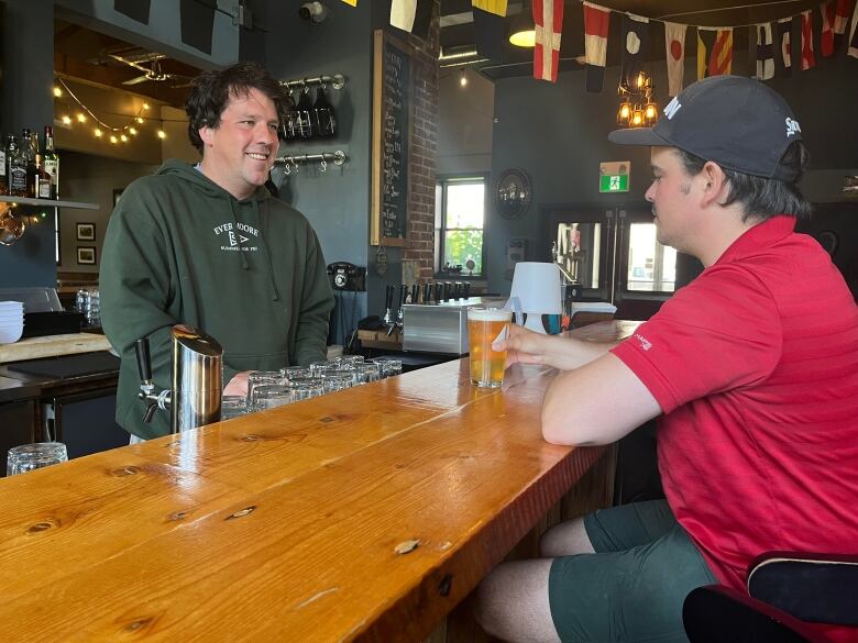 A man behind a bar smiles at a man holding a beer.