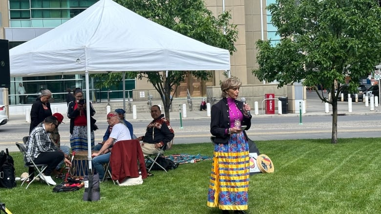 A woman in a ribbon skirt speaks in front of a circle of Indigenous drummers. 