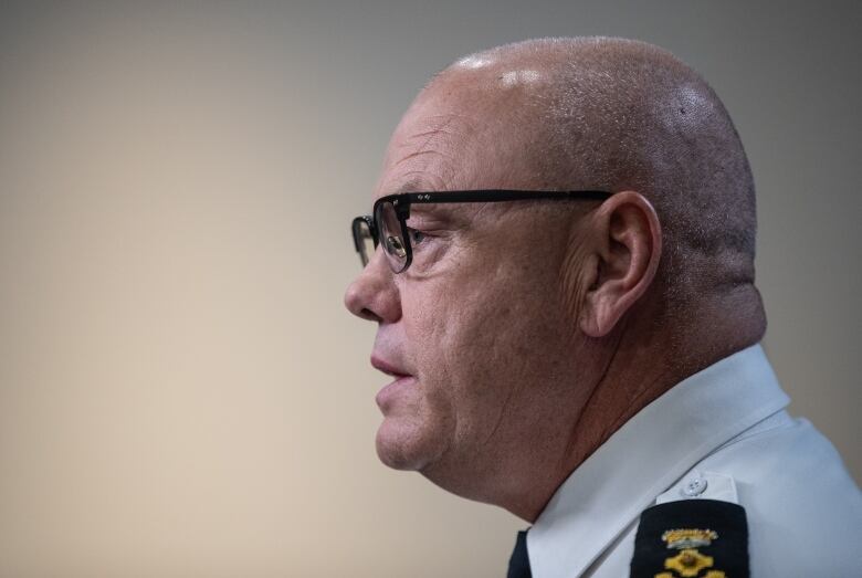 A bald man wearing glasses with a police patch on his shoulder is shown in front of a beige background.