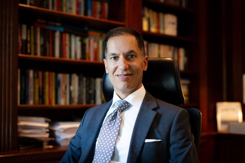 A man in a dark suit and a blue polka dot tie smiles while seated in front of a book shelf. 