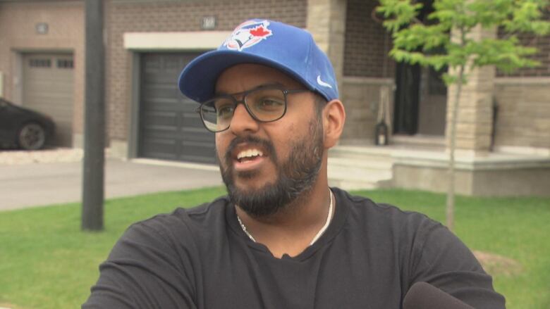 A man in a black shirt and blue cap stands in front of a house speaking.