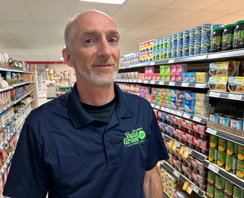 A man in a blue shirt stands in front of a shelf full of groceries.