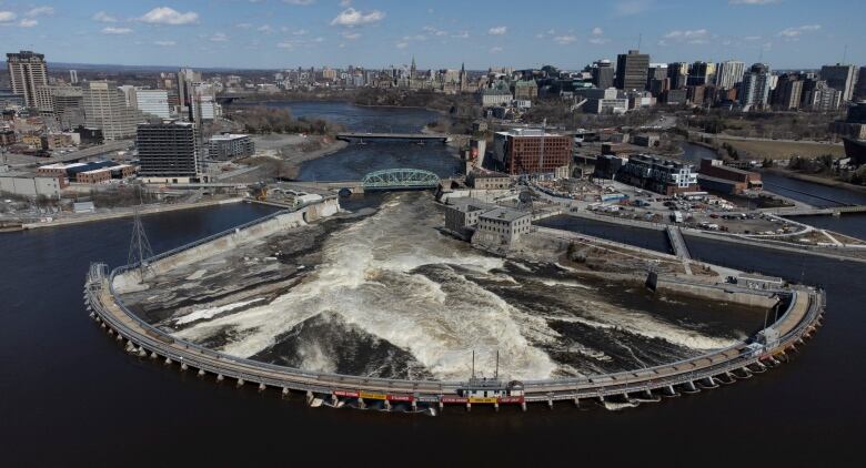 An aerial view of a half-moon shaped dam holding water back on the bottom of the photo, with the turbulent water seen in the middle of the photo, and a cityscape seen around it.