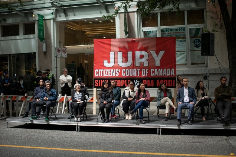 A group of people sit on an outdoor platform, with a sign behind them reading 'Jury Citizens' Court of Canada Sikh people vs PM Modi'.