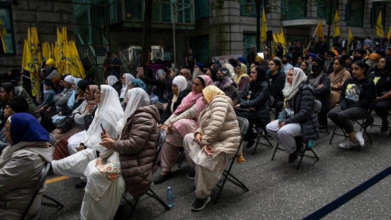 A number of Sikh people, many of whom are women, sit on chairs outdoors in a street.