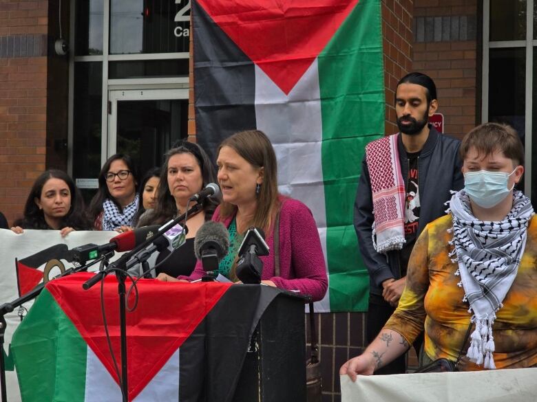 A woman speaks at a podium draped in a Palestinian flag with other protesters holding signs and flags in front of VPD HQ.