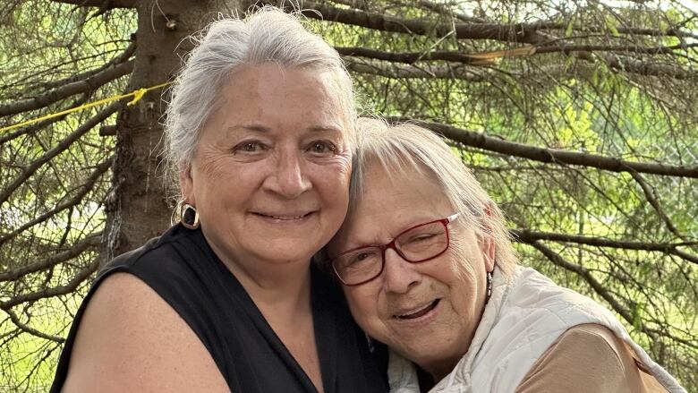 Two women with grey hair stand in a forest with their arms around each other.