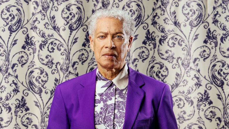 A Black man with white hair in front of a ornate background. He is wearing a purple blazer