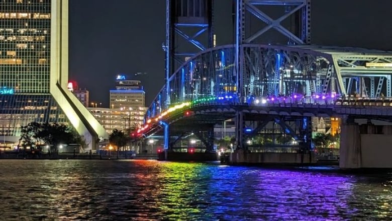 A night time view of a bridge spanning a river with a line of people holding red, orange, yellow, green, blue and purple lights that are reflecting on the dark water below. 