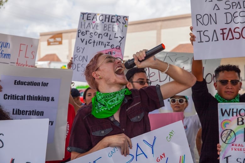 A person with pink-dyed hair, wearing glasses and a green handkerchief around his neck, shouts into a microphone while standing in front of a crowd of protesters. 