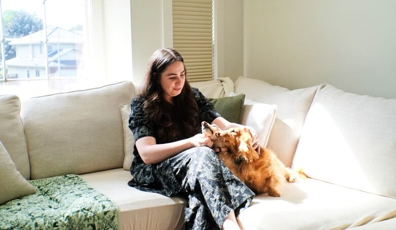 A woman in a patterned black outfit sits on a cream coloured couch with her Dachshund dogs. 