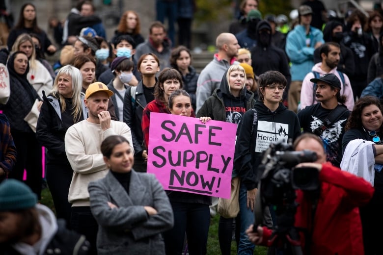 A group of people are seen at a protest. One of them holds a sign that reads 'Safe Supply Now!'