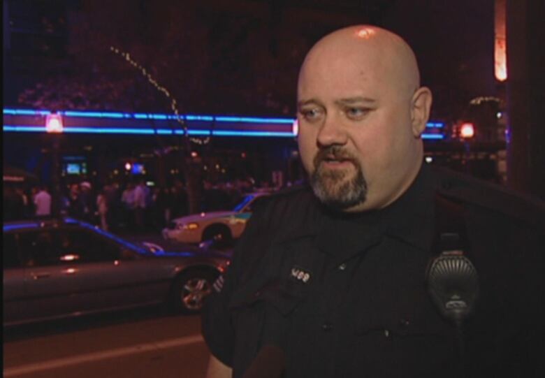 A bald white man wearing a police uniform speaks in front of a nightclub.