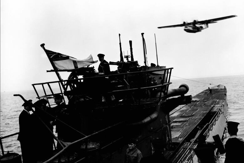 A black and white photo of a risen submarine with crew standing on it. A plane flies overhead.