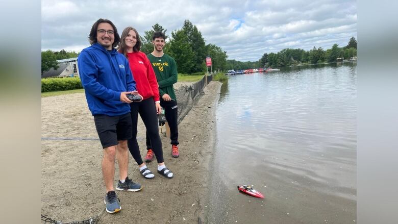 Three people stand at the bank of the beach holding controllers in front of a small remote-controlled boat in the water. 