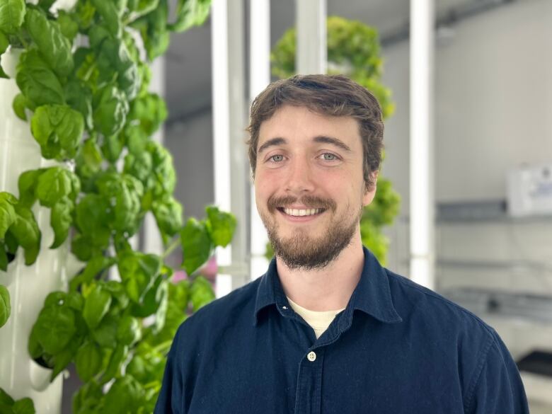 A smiling young man with brown hair and a short beard stands in a blue shirt in front of plant towers.