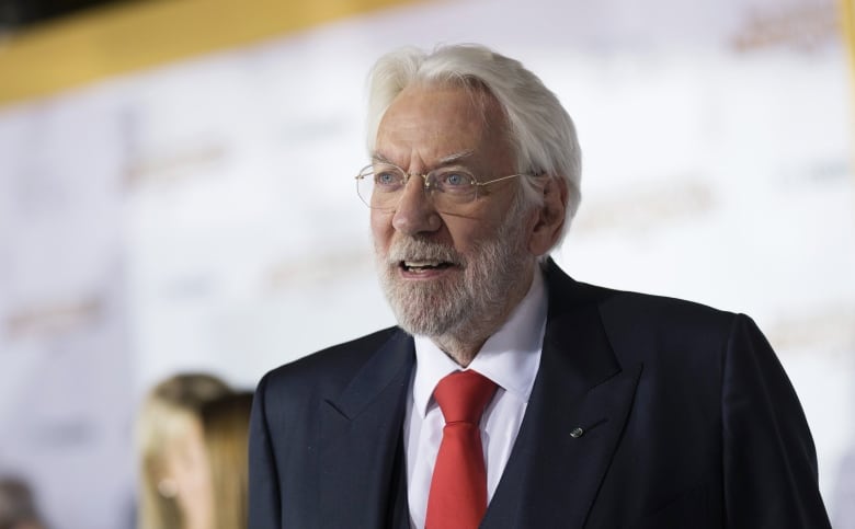 A grey-haired actor in a suit poses on a red carpet.