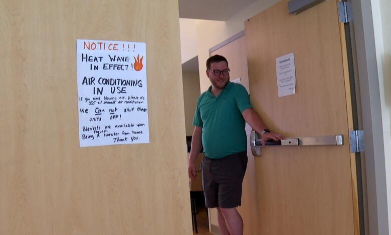 A man in a green polo shirt and shorts hold the door open. A sign on the door says Notice, Heat Wave in Effect.