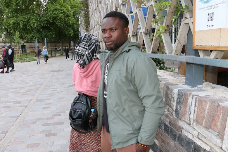 A man stands wearing a grey jacket. To his left is a woman with her back turned. 